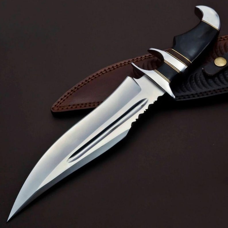 May include: A silver and black hunting knife with a serrated blade and a black handle with gold accents. The knife is resting on a brown leather sheath.