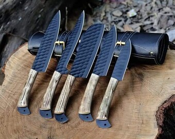 May include: A set of six black chef knives with textured blades and light wood handles. The knives are arranged on a wooden surface next to a black leather knife roll with gold buckles.