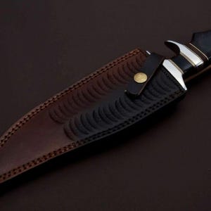 May include: A brown leather sheath with a black embossed pattern and a brass button. The sheath is designed to hold a knife.