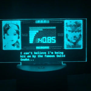 May include: A blue and white LED light with a pixelated image of a woman and a man. The text "MOBS", "MEMORY", "I can't believe I'm being hit on by the famous Solid Snake..." is displayed on the light.