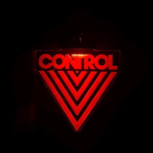 May include: A red, illuminated sign with the word "CONTROL" at the top. Below the text is a red, inverted triangle with three chevron-shaped lines. The sign is against a black background.