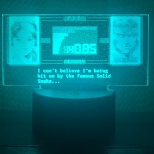 May include: A turquoise blue LED light with a clear acrylic panel displaying a pixelated image of two characters from a video game. The text on the panel reads "I can't believe I'm being hit on by the famous Solid Snake..."