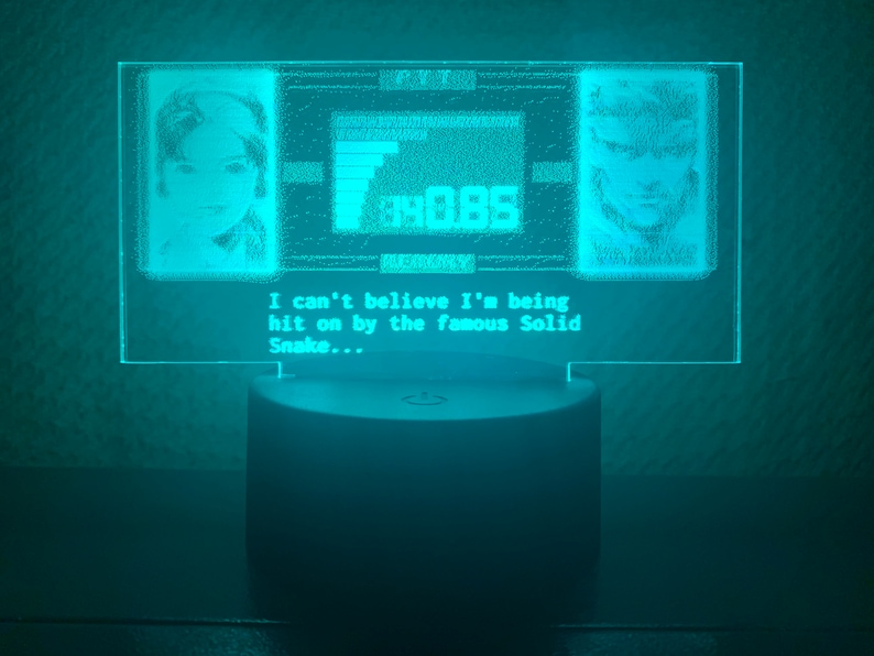 May include: A turquoise blue LED light with a clear acrylic panel displaying a pixelated image of two characters from a video game. The text on the panel reads "I can't believe I'm being hit on by the famous Solid Snake..."