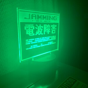 May include: A green LED sign with the text "JAMMING" and "電波障害" in Japanese. The sign is on a white base and is sitting on a dark surface. There is a "CAUTION" sign in the foreground.