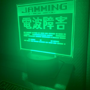 May include: A green LED light with a white base. The light features a pixelated design with the words "GAMING" and "電波障害" in Japanese. The light is on a dark surface with a "CAUTION" sign visible in the foreground.
