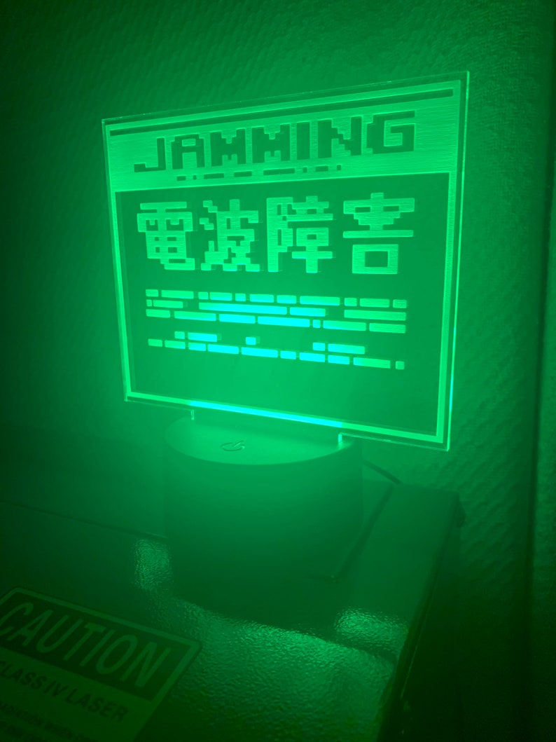 May include: A green LED light with a white base. The light features a pixelated design with the words "GAMING" and "電波障害" in Japanese. The light is on a dark surface with a "CAUTION" sign visible in the foreground.