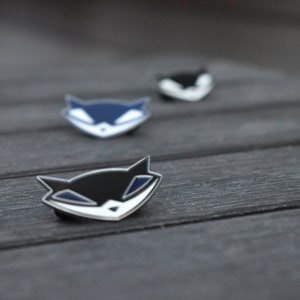 May include: Three enamel pins featuring a cartoon fox with a sly grin. The pins are in black, blue, and white.