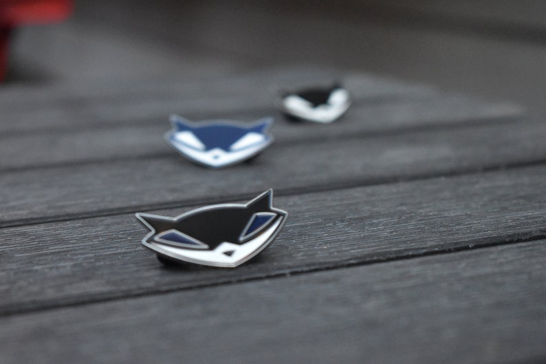 May include: Three enamel pins featuring a cartoon fox with a sly grin. The pins are in black, blue, and white.