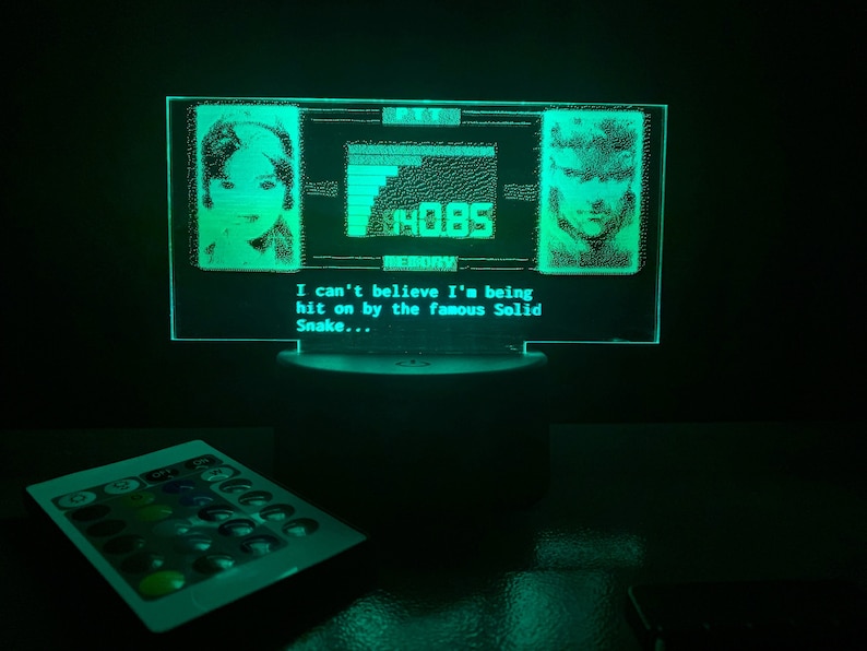 May include: A green LED light with a pixelated image of a woman and a man. The text "I can't believe I'm being hit on by the famous Solid Snake..." is displayed below the image. The light is on a black surface.
