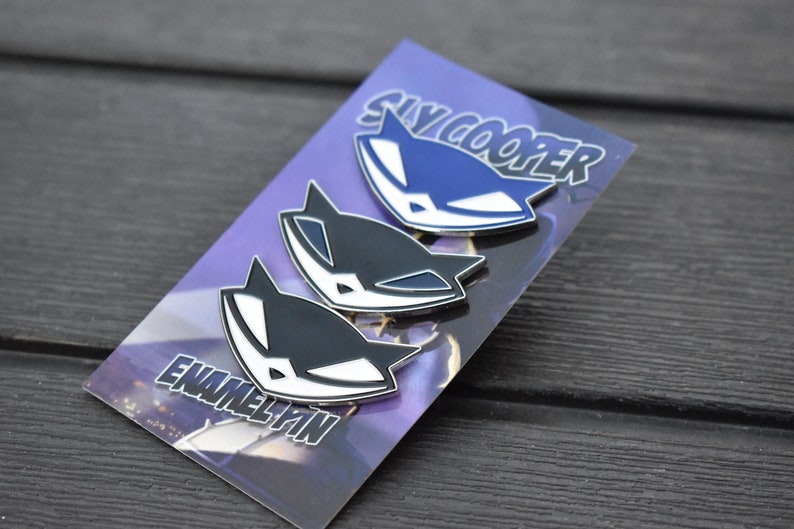 May include: Three enamel pins featuring the Sly Cooper logo. The pins are black and white, and blue and white. The pins are on a purple and black background with the text "Sly Cooper" and "Enamel Pin".