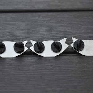 May include: Three silver metal pins with black rubber backs. Each pin features a stylized eye design.