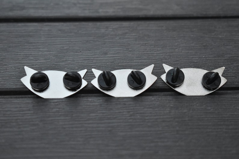 May include: Three silver metal pins with black rubber backs. Each pin features a stylized eye design.