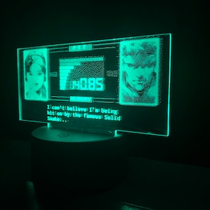 May include: A green LED light with a pixelated image of a woman and a man. The text "I can't believe I'm being hit on by the famous Solid Snake..." is displayed below the images.