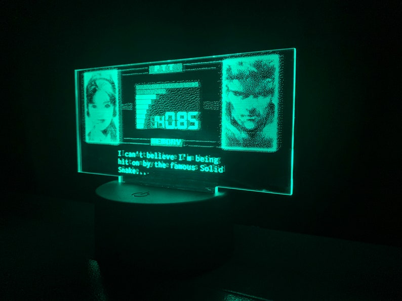 May include: A green LED light with a pixelated image of a woman and a man. The text "I can't believe I'm being hit on by the famous Solid Snake..." is displayed below the images.