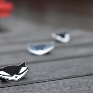 May include: Three enamel pins featuring a black and white cartoon fox with a menacing expression. The pins are arranged in a row on a wooden surface.