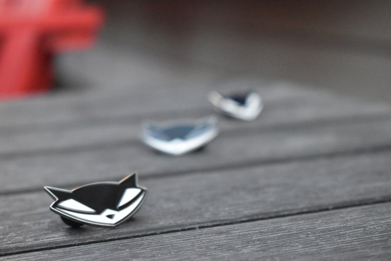 May include: Three enamel pins featuring a black and white cartoon fox with a menacing expression. The pins are arranged in a row on a wooden surface.