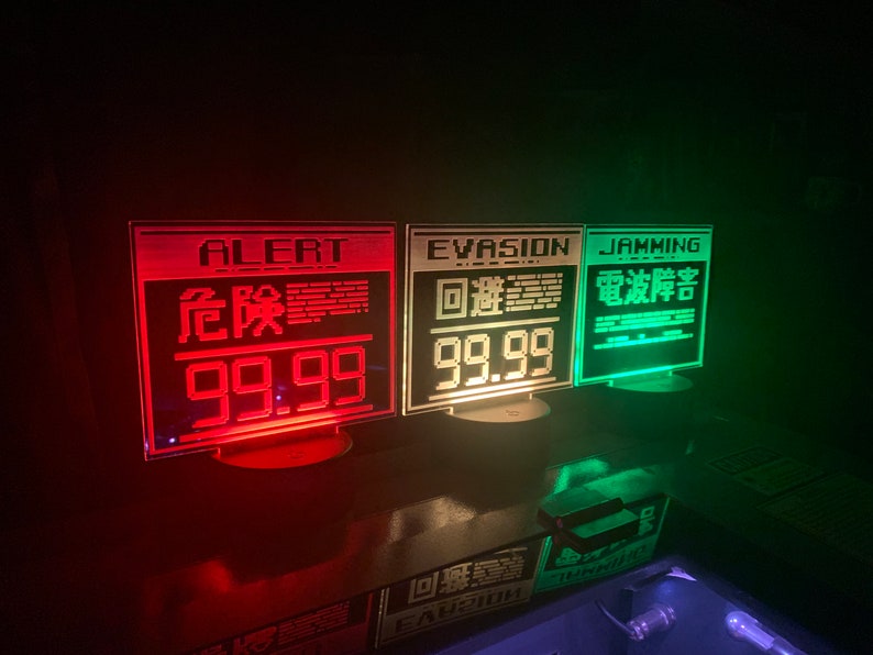May include: Three red, white, and green illuminated signs with black text. The first sign reads "ALERT" in white, "危险" in red, and "99.99" in red. The second sign reads "EVASION" in white, "迴避" in white, and "99.99" in white. The third sign reads "JAMMING" in white, "電波干渉" in white, and "99.99" in white.