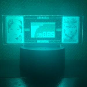 May include: A teal blue acrylic lamp with a rectangular base featuring two etched portraits and a small rectangular design in the center. The lamp is on a dark surface.