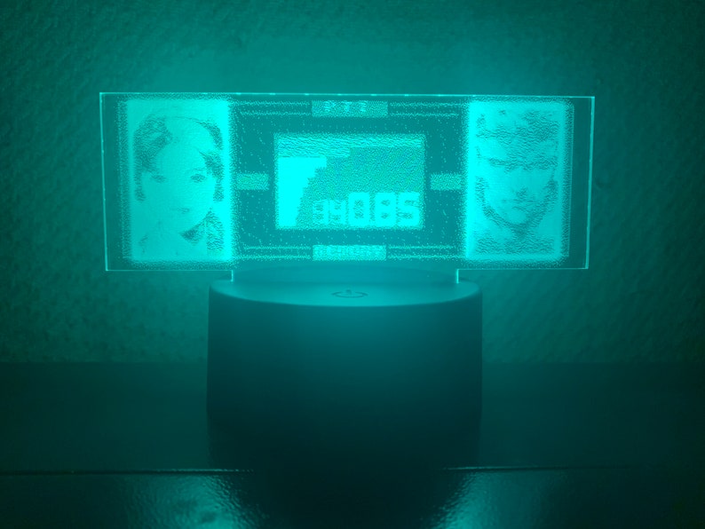 May include: A teal blue acrylic lamp with a rectangular base featuring two etched portraits and a small rectangular design in the center. The lamp is on a dark surface.