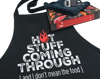 May include: Black apron with a white graphic that says "HOT STUFF COMING THROUGH {and I don't mean the food}". The apron has a pocket on the left side. A small box with a blue dish towel and a bottle opener is in the background.