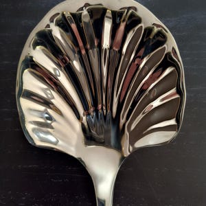 May include: A silver-colored serving spoon with a fan-shaped head. The spoon's head has a fluted design, and the handle is smooth and slender. The spoon is likely used for serving food.