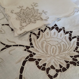 May include: Close-up of a cream-colored linen tablecloth and a matching napkin, both featuring intricate floral embroidery. The tablecloth has a large, openwork floral design with a scalloped edge. The napkin has a smaller, similar floral design.