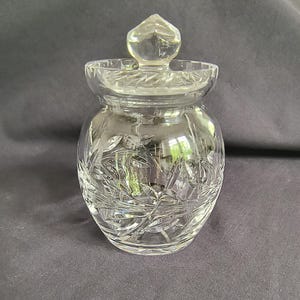 May include: A clear, cut-glass jar with a lid. The jar has a rounded body and a lid with a pointed finial. The glass is intricately cut with a leaf and floral pattern. The jar is likely used for storage or display.