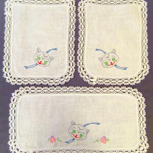 May include: Set of three white linen placemats with a crocheted lace border. Each placemat features an embroidered gray cat face with green eyes, a red mouth, and a blue ribbon. The bottom placemat has embroidered pink flowers.