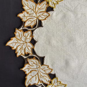 May include: A beige linen placemat featuring a scalloped edge decorated with embroidered maple leaves in shades of white and golden yellow, outlined with dark brown thread, creating a detailed botanical border design.
