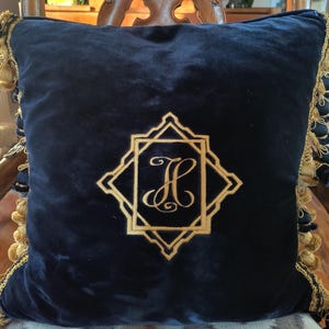 May include: Navy blue velvet throw pillow with gold embroidered initial "H" inside a geometric frame. The pillow has gold fringe trim around the edges. The pillow is square in shape.