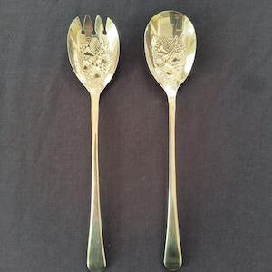 May include: A pair of gold-colored serving utensils, a fork and a spoon, with ornate embossed designs on the bowl. The fork has three tines. The spoon has a rounded bowl. Both have long, slender handles.