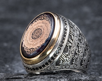 May include: A silver ring with an oval-shaped black stone set in a gold bezel. The stone is engraved with intricate Arabic calligraphy. The ring band is decorated with a detailed floral pattern.