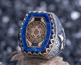 May include: A silver ring with a blue stone set in a gold-toned, ornate design. The stone features a six-pointed star and Arabic script.