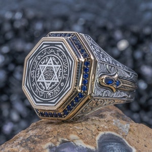May include: A silver ring with a gold octagonal centre featuring a six-pointed star and Arabic script. The ring is decorated with blue gemstones.