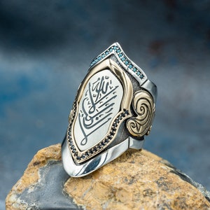May include: A silver and gold ring with a shield-shaped design. The shield features black and blue gemstones and Arabic calligraphy.