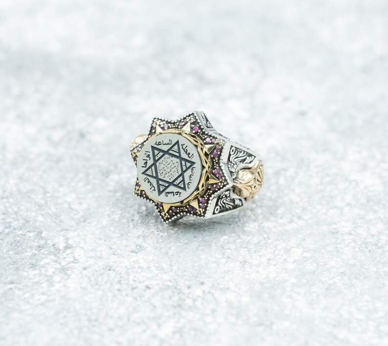 Puede incluir: Un anillo de plata con un dise&ntilde;o de estrella de David y escritura &aacute;rabe. El anillo est&aacute; engastado con peque&ntilde;as piedras preciosas rosas alrededor de la estrella.