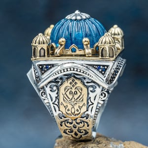 May include: A silver and gold ring featuring a blue dome-shaped stone and intricate details. The ring is designed in the style of a mosque.