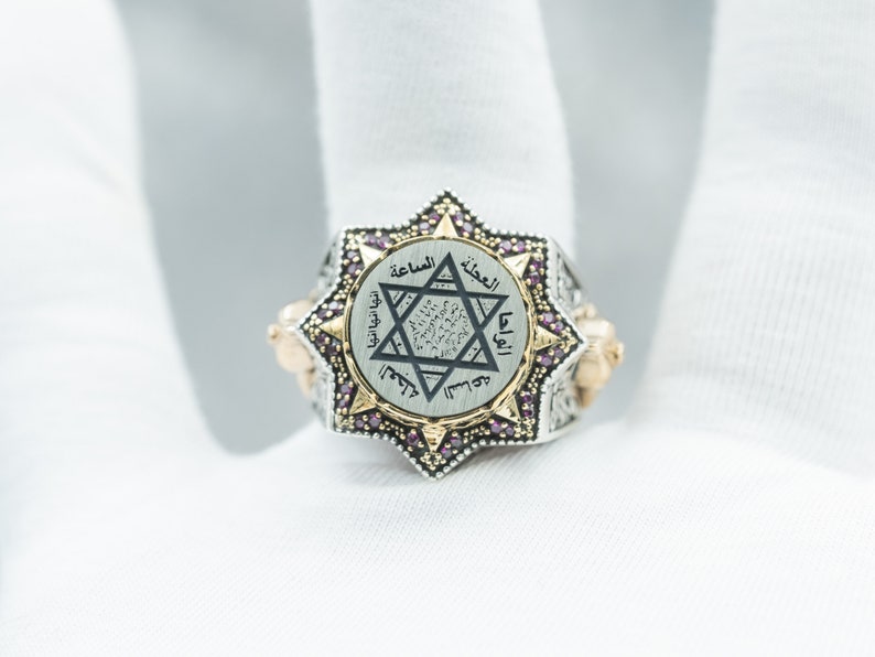 Puede incluir: Un anillo de plata y oro con un dise&ntilde;o de estrella de David y escritura &aacute;rabe. El anillo est&aacute; engastado con peque&ntilde;as piedras preciosas rosas.