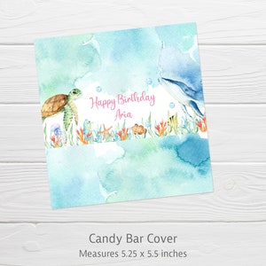 May include: A blue and green watercolor candy bar cover with a whale, sea turtle, and other sea creatures. The text reads "Happy Birthday Aria". The cover measures 5.25 x 5.5 inches.
