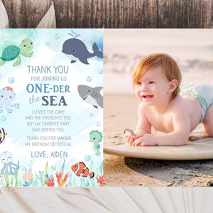 A baby is lying on a surfboard on a beach, surrounded by ocean-themed decorations, with a "Thank you for joining us ONE-DER the sea" card featuring a baby and sea animals on the left side.
