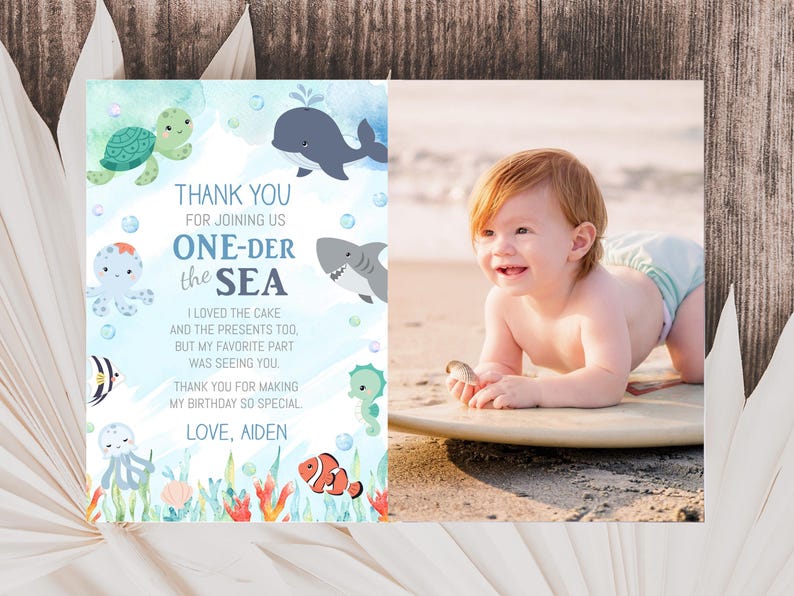 A baby is lying on a surfboard on a beach, surrounded by ocean-themed decorations, with a "Thank you for joining us ONE-DER the sea" card featuring a baby and sea animals on the left side.