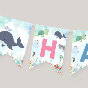 Può includere: Una ghirlanda decorativa con illustrazioni ad acquerello di creature marine tra cui una balena, una tartaruga, uno squalo e un polpo. I gagliardetti mostrano le lettere H e A in rosa e blu, su uno sfondo azzurro con alghe marine.
