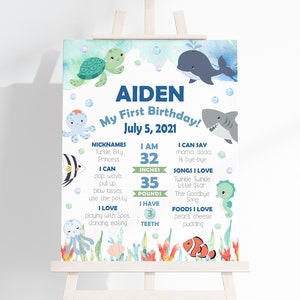 Puede incluir: Un cartel de primer cumplea&ntilde;os de estilo acuarela azul y verde con tem&aacute;tica de criaturas marinas. El cartel presenta el nombre Aiden, la fecha 5 de julio de 2021, y detalles sobre el primer a&ntilde;o del ni&ntilde;o, incluyendo su altura, peso, alimentos y canciones favoritos.