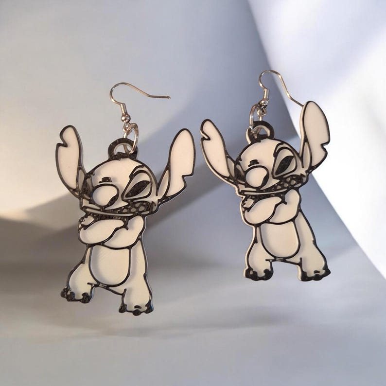 May include: A pair of dangle earrings featuring a white, cartoon-style character with large ears and crossed arms, outlined in bold black lines. The earrings are suspended from silver-toned metal hooks, showcasing a flat, graphic design.