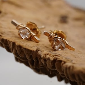 May include: A pair of gold earrings with clear quartz stones wrapped in wire. The earrings are on a wooden surface.