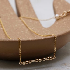 May include: A gold chain necklace with a bar of small gold beads. The necklace is displayed on a brown textured surface.