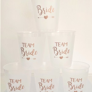 May include: Set of six clear plastic cups with gold lettering. The cups have the words "Bride" and "Team Bride" printed on them.