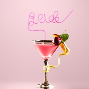 May include: A pink cocktail in a martini glass with a pink straw that says "bride" on it. The drink is garnished with a cherry, a slice of mango, and a sprig of mint.