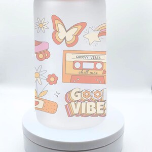 May include: A frosted glass water bottle with a bamboo lid and a straw. The bottle has a retro design with a butterfly, a cassette tape, a roller skate, and the words "Good Vibes" and "Groovy Vibes chill mix".