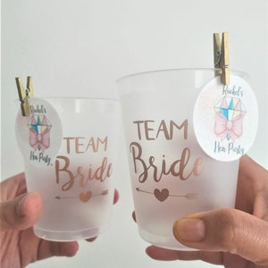 May include: Two frosted plastic cups with the text "Team Bride" in gold lettering. Each cup has a small pink and white sticker with a bow and the text "Rachel's Hen Party".